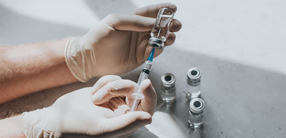 A hand wearing latex gloves hold a phial of clear liquid and a syringe. The medical professional uses the syringe to extract the medicine from the glass bottle. Conceptual with space for copy.