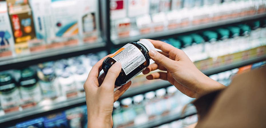 Over the shoulder view of young Asian woman browsing through medical products and reading the label on a bottle of medicine in front of the shelves in a pharmacy
